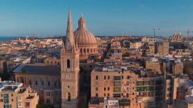 Gün batımında Valletta, Malta 'nın üzerinde bazilika kubbesi, profesyonel katedral kulesi, dar sokaklar, çatı terasları, mavi ve yeşil balkonlar ve ufukta liman vinçleri görünüyor..