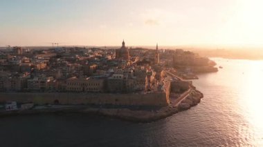 Gün batımında Valletta, Malta havadan bakıldığında kireçtaşı çatılar, bazilika kubbesi, St. Pauls Pro Katedrali kulesi, kıyı tahkimatları, deniz kıyısı, yol ve liman tekneleri görülüyor..