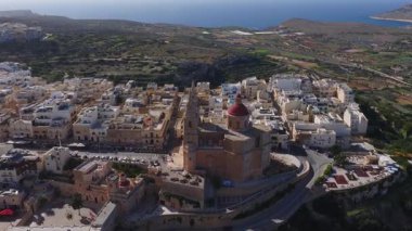 Hava aracı taraması, Mellieha, Malta 'da kırmızı kubbeli bir kilise, düz çatı blokları, teraslı alanlar, kıyı yolları ve gün ışığında uzak Comino ve Gozo' yu gösteriyor..