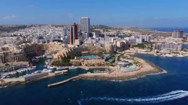 Sliema, Malta 'nın gün ortası görüntüsünde Tigne Point sahili, marina, kırmızı fener, lido havuzu, yüksek binalar, vinçler ve canlı panoramik renkte bir motorlu tekne görülüyor..