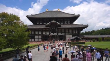 Japonya, Nara 'daki Todai ji' deki Daibutsuden 'ın gündüz görüşü. Ziyaretçiler taş gezinti alanında şemsiyelerle yürürler. Tahta ön cephe, fayanslı çatı, görünür altın şibi..