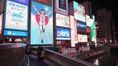 Osaka 'daki Dotonbori Kanalı' nda turistler yürürken ve fotoğraf çekerken gece sahnesi. Glico koşucusu, Chintai, SAMTY ve 100. yıldönümü işaretleri parlıyor ve suya yansıyor.