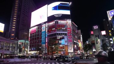 Shibuya 'nın Tokyo' daki geçişinin geniş açılı görüntüsü. Aydınlatılmış mağaza vitrinleri, UNIQLO tabelası, Samsung ekranı, ve 109 kule çerçeve araba, otobüs, bisiklet ve yayalar.