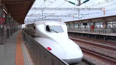 Shin Yokohama 'da 14. peronda beyaz bir N700 Shinkansen bekliyor. Bir kondüktör pencereden sarkar. Bulutlu gün ışığı, düşük açı çizgileri, katenareri sıraları çekiliyor.