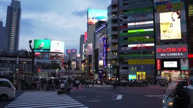 Tokyo 'daki Shibuya Scramble' da sokak manzarası yayalar, arabalar, otobüsler, kamyonlar, bisikletçiler ve BIC CAMERA, Shibuya 109 ve LED reklamları arasında gidip gelen kartları gösteriyor..