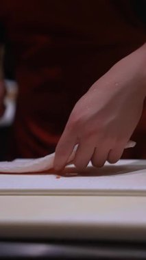 Yakından bakınca şefin beyaz tahtada sashimi bıçağıyla ıslak beyaz balığı dilimlediğini görüyoruz. Deri ve pullar ince kesikler olarak gösteriliyor. Ilık alçak ışık, sığ derinlik.