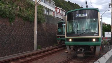 İki yeşil ve krem rengi Enoden treni Japonya 'nın Kamakura şehrinde dar bir kıyı şeridinde buluşuyor. El bilgisayarı gündüz görüntüsü platform düzeyi çekimleri ile geniş ve kapalı olarak görüntülenir.
