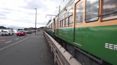 Antika yeşil ve krem rengi Enoden treni 305 ve 355 numaralı otoyollar ve Kamakura 'da bir gezinti güvertesi üzerinden Enoshima ve deniz feneriyle birlikte geçmektedir..