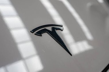 Tesla T amblemini kapalı bir garajdaki parlak beyaz panele kapatın. Yumuşak pencere yansımaları yüzey boyunca çizgiler, yüzeyin sığ derinliği metalik hatları vurgular.