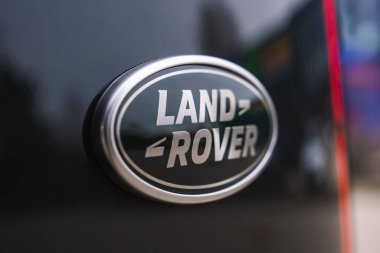 Yüzeyin sığ derinliği parlak siyah boyada gümüş ve koyu yeşil Land Rover rozeti yumuşak ışıkta parlak harfler, atölye yansımaları gösteriyor..