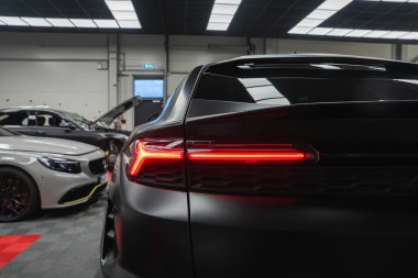 Siyah bir Lamborghini Urus SE arka üç çeyrek görüş, LED stop lambası yanıyor. Beyaz bir Mercedes Benz AMG coupe parlak bir servis atölyesinde açık bir kaputun yanında oturuyor..