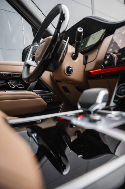 Range Rover otobiyografisinin iç mekânında bronz deri, dijital küme, metal kürekler ve yumuşak endüstriyel ışıklandırmalı prestijli bir atölyede ışıltılı konsolda vites seçici görülüyor..