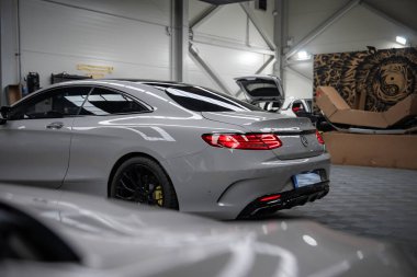 Modern bir atölyeye park edilmiş gümüş renkli bir Mercedes Benz AMG Coupe, LED stop lambaları açık, dört egzoz uçları görünür, siyah tekerlekler ayarlanmış. Açık başlıklar, aslan duvar resmi ve parlak ışıklandırma ayarlama önerir.