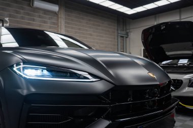 Mat siyah Lamborghini Urus 'a yakın modern bir atölyede parlayan Y şeklindeki LED farlı, Mercedes Benz AMG Coupe kapüşonlu, endüstriyel ışıklandırmalı.