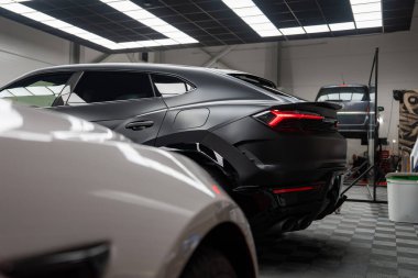 Siyah bir Lamborghini Urus ışıklı bir atölyede duruyor, parlak arka farları görülebiliyor. Bir Mercedes AMG ve yeni bir Range Rover serviste, biri asansörde, etrafta aletler var..