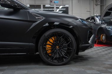 Yakın plan, mat siyah Lamborghini Urus SE önü, siyah çoklu konuşma tekerleği, sarı kalibre arması gösteriyor. Parlak atölyede Range Rover ve AMG coupe bakım altında.