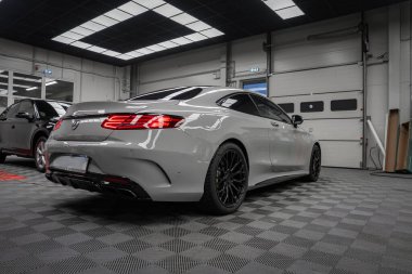 Beyaz Mercedes Benz AMG Coupe bir servis hangarında oturuyor, arka üç çeyreklik, LED stop lambaları, siyah jantlar, dört tekerlek egzozları, dama döşemeleri, parlak şebeke ışıkları, yakındaki SUV 'ler.