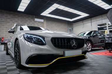 Düşük açılı geniş açı, parlak tavan panelleri altında Lamborghini Urus ve Range Rover ile kareli bir atölye zemininde park etmiş beyaz bir Mercedes AMG coupe gösteriyor..