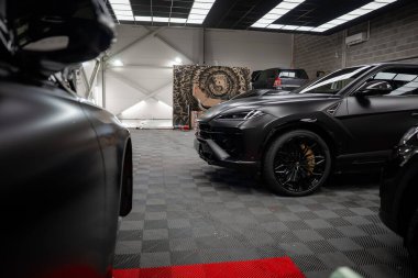 Matte Black Lamborghini Urus, şebeke ışıkları altında bronz tekerleklerle sağda duruyor. Range Rover ve Mercedes coupe gri zeminde kamuflaj bölmesi ve armanın arkasında oturuyorlar..