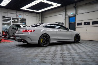 Siyah tekerlekleri ve sarı kalibreli gümüş bir Mercedes AMG coupe, lüks bir SUV ve bakım altında arabalar, LED paneller ve görünür dama zemin ile parlak bir atölyede oturuyor.