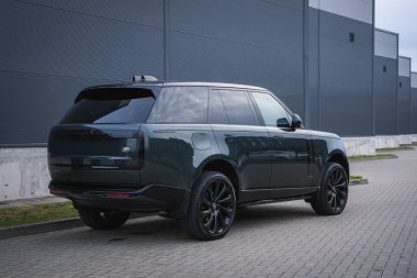 Siyah tekerlekleri olan koyu yeşil Range Rover gri metal bir binanın yanındaki asfalt servis şeridinde duruyor. Serin gün ışığında alçak arka üç çeyrek manzara temiz çizgiler gösterir..