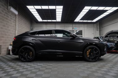 Bir mat siyah Lamborghini Urus SE modern bir atölyede, kareli zemin, şebeke ışıkları, Y konuşmalı tekerlekler ve altın kalibreli arabalar yakınlarda hizmet vermekteyken.