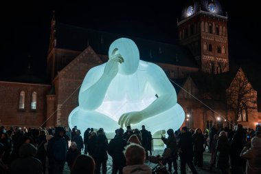 Gecenin bir yarısı Riga Eski Şehir meydanında Riga Katedrali arkasında Riga Katedrali ve ziyaretçilerinin toplandığı açık mavi beyazla aydınlatılmış saydam bir şişme figür oturuyor..