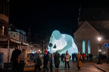 Riga, Letonya 'daki gece sahnesinde ziyaretçiler St. Peter Kilisesi' nde toplanıyor. Cyan beyazı bir şişirilebilir, Staro Riga 2025 sırasında insanların fotoğraf çekmesi ve etkileşime girmesi için tuğla duvarların ve kemerli pencerelerin önünde diz çöker..