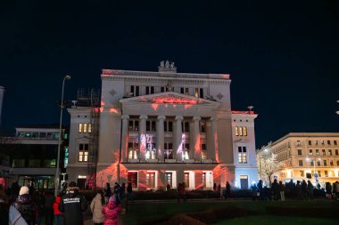 Riga 'daki Letonya Ulusal Operası, kırmızı ve beyaz ışığın ön cepheyi haritaladığı gece parlıyor. Kışlık ceketli kalabalık, tarihi mimarinin ortasında Staro Riga 2025 'i izliyor..