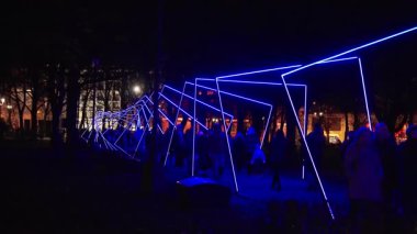 Riga, Letonya 'da gece festivali, mavi ve kırmızı renkli açısal neon çerçeveler gösteriyor, kalabalıklar tarihi cephelerle dolu ağaçların arasında yürürken siluetler dökülüyor..
