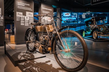 Pirinç tankı ve korunmasız motoru olan klasik bir motosiklet, bir otomobil müzesinde yükseltilmiş bir platformda, Baltık dilinde pankartlar, havalı ışıklandırma altında eski bir araba,.