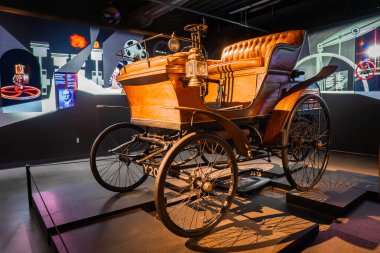 Yenilenmiş atsız bir araba kapalı bir otomobil müzesinde bir platformda duruyor, verniklenmiş ahşap, pirinç fenerler, tekerlekli sandalyeler, ve sıcak, yönlü ışık tarafından aydınlatılmış deri koltuklar..