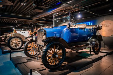 1900 'lerin başında, parlak mavi renkli, pirinçten farları olan bir turne arabası, dramatik ışıkların altında, bir müzenin platformunda, üzerinde numaralandırılmış bir yarış stili bulunur..