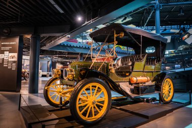 1900 'lü yılların başlarında açık bir turne arabası, spot ışıkları altında bir platforma oturur. Pirinç lambalar ve krank görünür, sarı ahşap tekerlekler, deri koltuklar ve tepe örtüsü görünür..