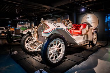 Beyaz ve kırmızı 20. yüzyılın başlarında 10 numaralı yarış arabası yakınlarda yeşil Ford Model T ve pirinç detaylarıyla bir otomobil müzesinde küratörlü aydınlatma altında duruyor..