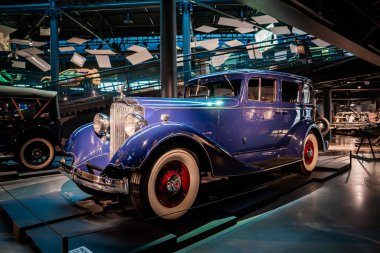 1930 'ların beyaz duvarları ve kırmızı kablo tekerlekleri olan mavi lüks bir sedan modern bir otomobil müzesinde spot ışıkları altında duruyor, yerleri cilalıyor ve Art Deco çizgilerini ön plana çıkarıyor..