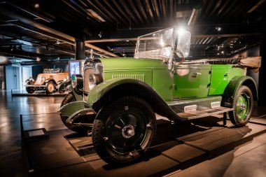 Loş bir otomobil müzesi içinde, parlak yeşil, üstü açık, klasik bir araba, bariyerler, cilalı zemin ve sıcak yönlü ışık.