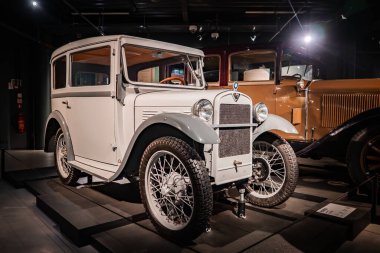 Bir otomobil müzesinin yükseltilmiş platformunda beyaz bir mikroaraba duruyor, spot ışıklarıyla aydınlatılıyor, tel teller, ön menteşeler, küçük bir ızgara rozeti ve arkasında bir hardal arabası var..