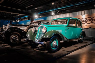 1930 model deniz mavisi bir sedan, lüks bir arabanın yanındaki bir otomobil müzesinde sıcak ışıkların altında durur. Otomobil Birliği halkaları ve 1932 Sergiyi krom kaplama ve konuşmalı tekerleklerle işaretle.