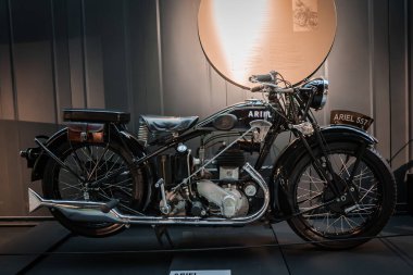 Korunmuş bir ARIEL 557 motosiklet bir müze platformunda, beyaz çizgili siyah bir tank, deri eyer, alet çantası, koyu paneller ve dairesel bir spot ışığı ile yan yana durur..