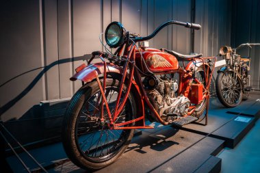 20. yüzyılın başlarında kırmızı bir motosiklet loş bir otomobil müzesinde yükseltilmiş bir platformda duruyor. Açığa çıkan çift motorlu, telli tekerlek, el vitesi ve altın çizgili tank görülebilir..
