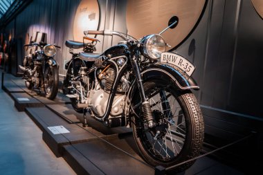 Cilalı bir BMW R 35 kapalı bir otomobil müzesinde yükseltilmiş bir platformda duruyor, arkasında başka bir klasik bisiklet, yumuşak galeri ışıklandırması, plaketler ve krom aksanlar var..