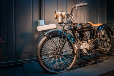 Antika bir motosiklet müzede duruyor, pirinç asetilen far, deri eyer, bozuk tekerlekler, açık motor, kemer sürücüsü ve kontrollü ışık altında el düdüğü..