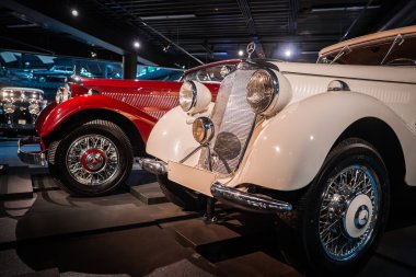 Yenilenmiş iki eski araba, müze ışıklandırmasının altındaki platformlarda duruyor, krem rengi bir Mercedes Benz roadster ve koyu kırmızı klasik, düşük açılı, krom ve detaylar..