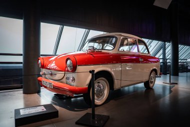 İki tonluk kırmızı ve krem renkli bir Trabant mikro arabası, modern bir salonda sergi ışıkları altında, ayakta duran ve bir pankartla etiketlenmiş, kapalı bir yerde duruyor..