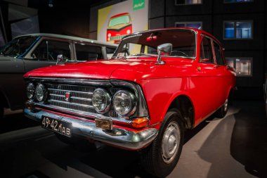 Parlak kırmızı Sovyet dönemi bir sedan müze ışıklandırması, krom parıltılar, siyah plaka 49 LAV, kutu gövde ve jant kapakları altında oturur, klasik araçlar ve duvar grafikleriyle çevrilidir.