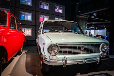 Açık yeşil bir Lada VAZ 2101 kapalı bir otomobil müzesinde parlak kırmızı klasik bir arabanın yanında duruyor. StandWithUkrayna çıkartması modern ışıklandırma altında ön camı işaretliyor.