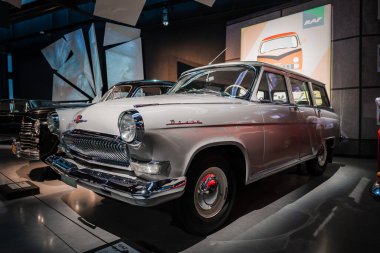 Beyaz bir GAZ 21 Volga vagonu bir otomobil müzesinde duruyor, krom ızgara ve yuvarlak lambalar parıldıyor, RAF logosu posterleri arkada oturuyor, yerleri cilalıyor ve dikkatli ışıklandırma drama ekliyor..