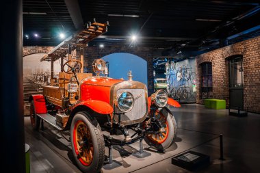 20. yüzyılın başlarında kırmızı çamurlukları, pirinç tesisatları ve ahşap merdiveni olan bir itfaiye arabası, tuğla duvarlar ve endüstriyel sergilerle çerçevelenmiş sıcak müze ışıklarının altında duruyor..