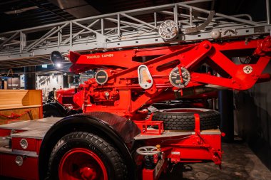 Bir otomobil müzesinin yakın görüntüsünde geniş merdiveni, vinç tekerlekleri, etiketli parçaları ve sergi ışıkları altında el kolları olan klasik kırmızı bir Magirus merdiven kamyonu görülüyor..